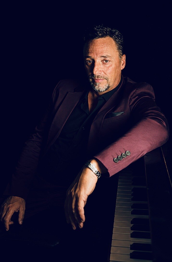 Jack Rivers beside a piano in a burgundy suit